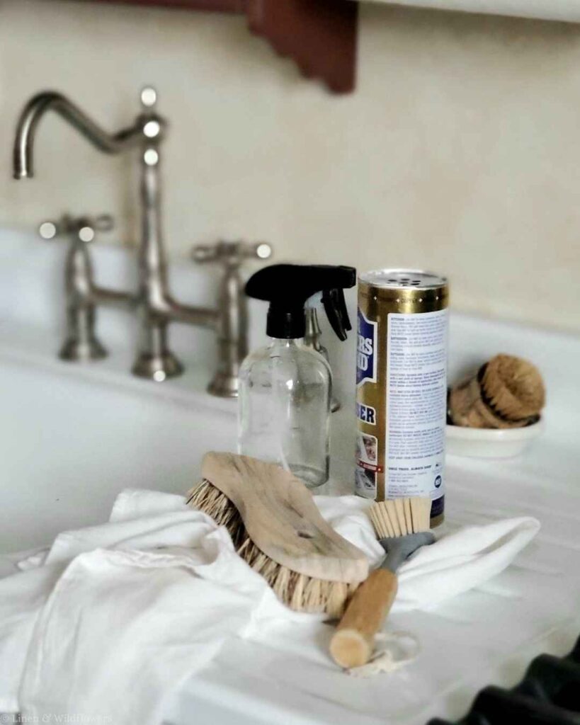 Bar Keepers Friend, glass spray bottle, and scrub brushes on a white farmhouse sink for easy kitchen cleaning.