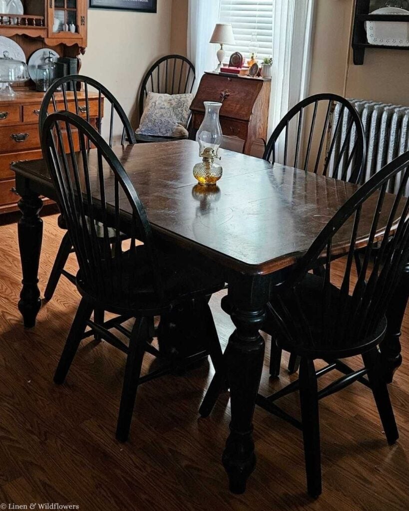 Vintage dining room with a dark wood table, black Windsor chairs, an amber oil lamp, and an antique secretary desk.