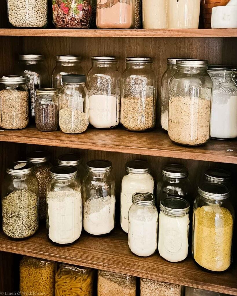 Pantry organization with glass jars of rice, flour, grains, and pasta stored in bulk on wooden shelves.