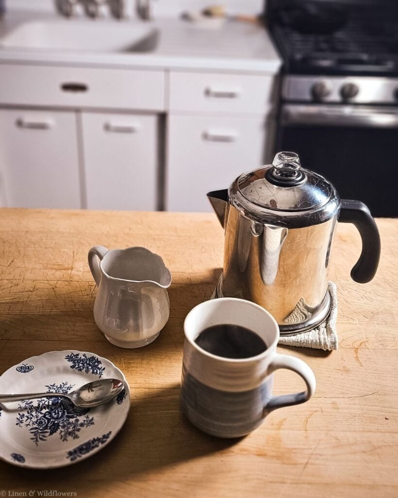 Clean stainless steel percolator beside a mug of hot black coffee, ironstone creamer and blue transferware saucer.
