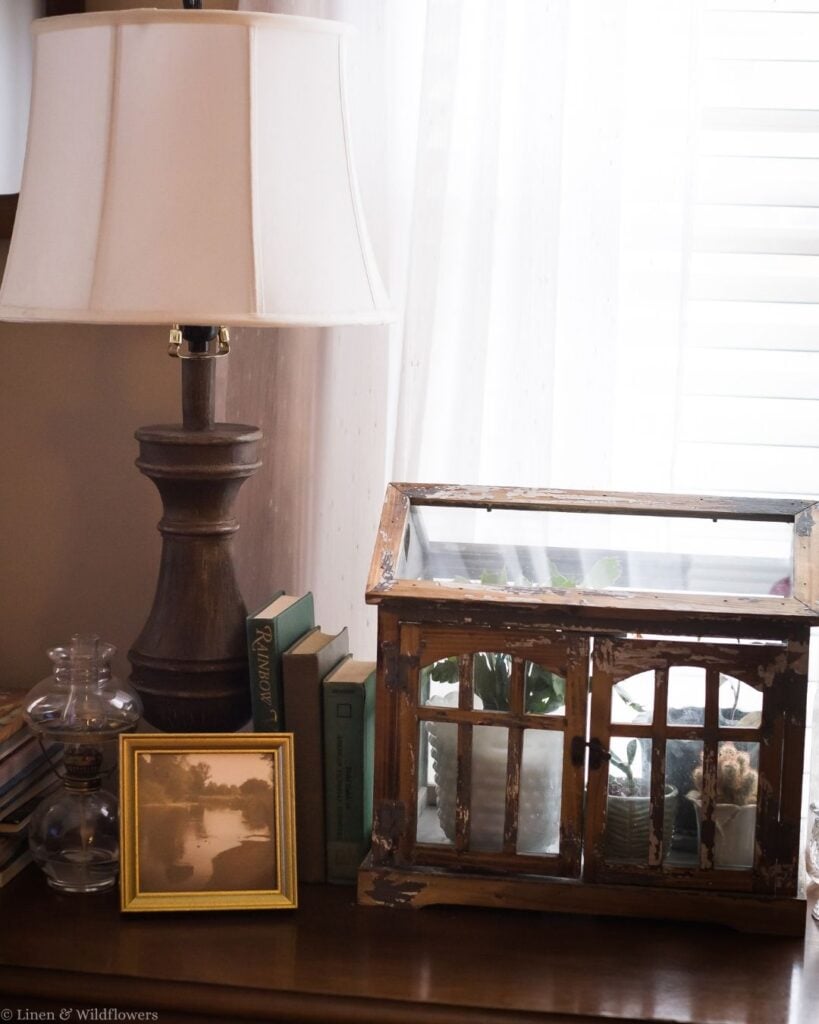 Vintage vignette with a table lamp, antique books, oil lamp, gold frame, and distressed wooden greenhouse planter.