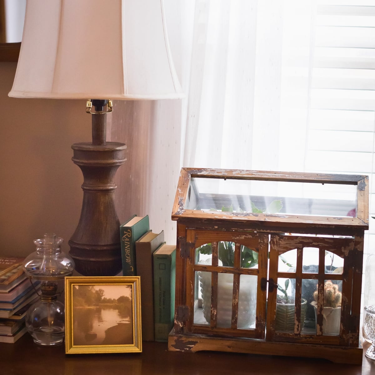 Vintage vignette with a table lamp, antique books, oil lamp, gold frame, and distressed wooden greenhouse planter.