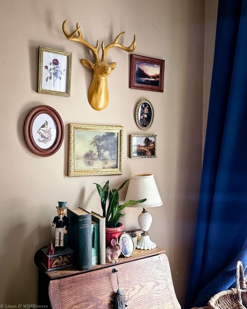 Vintage gallery wall with a gold deer head and thrifted frames above an antique oak secretary desk with books.