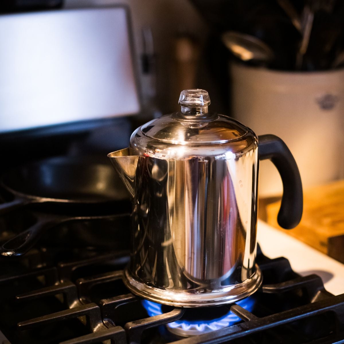 Polished stainless steel stovetop percolator with a glass knob lid on a gas stove beside a cast iron skillet.