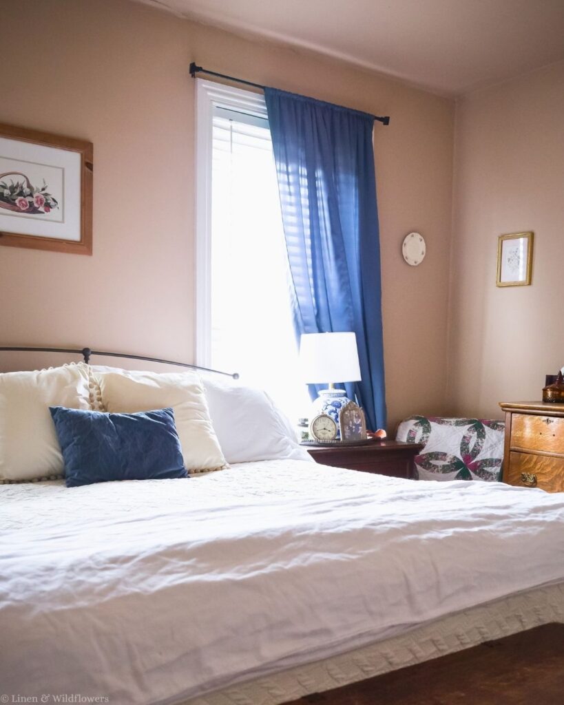 Bed made with white linens over a mattress protector pad in a vintage-inspired bedroom with a blue accent pillow.