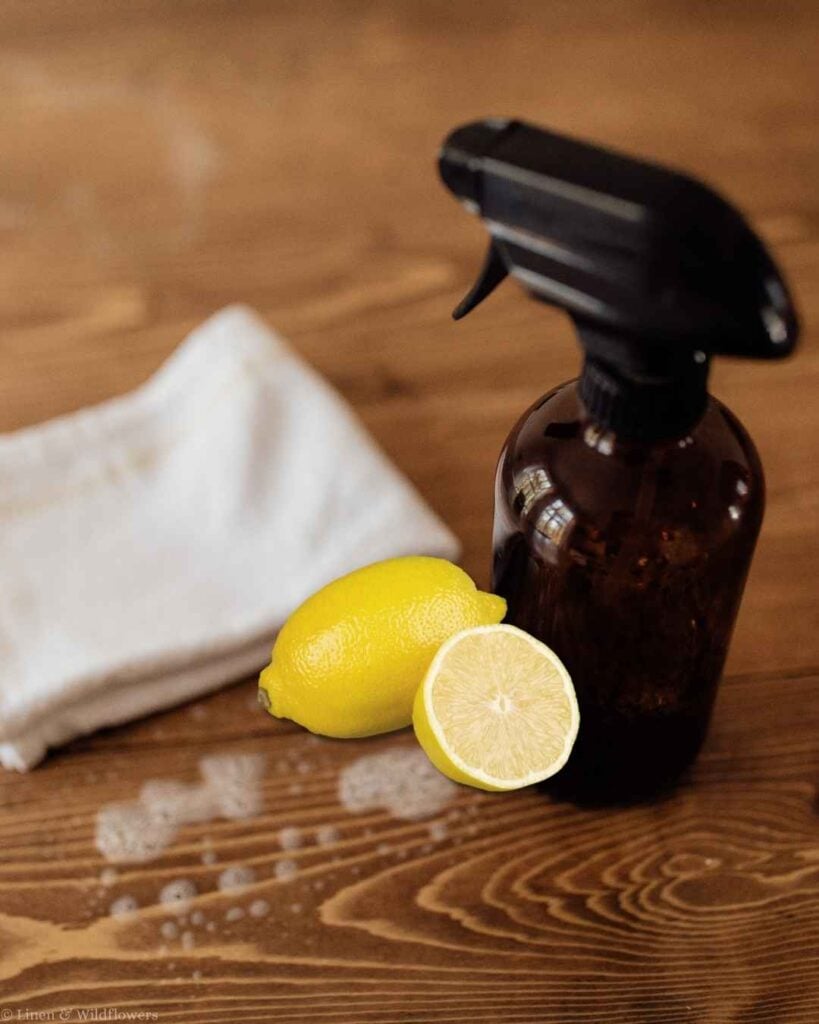Amber glass spray bottle with fresh lemons and a white cloth on a wood table for a homemade lemon cleaning spray.