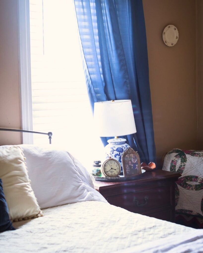 Cozy bedroom with a mattress protector pad under white bedding, a blue and white lamp, and blue curtains.