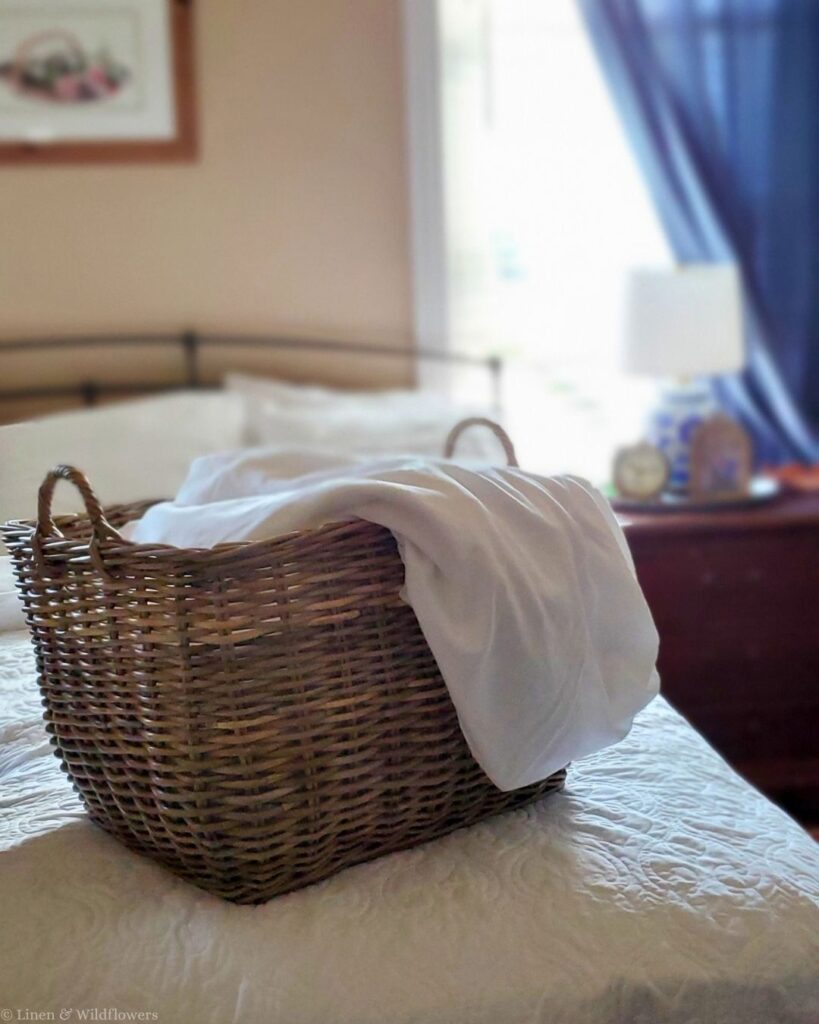 Wicker laundry basket with clean white sheets on a bed, ready to wash with a mattress protector pad.