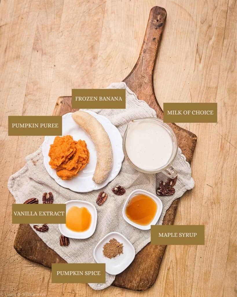 Pumpkin pie smoothie ingredients on a wooden board: pumpkin puree, frozen banana, milk, maple syrup, vanilla extract and pumpkin spice.