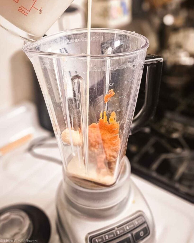 Pouring milk into a KitchenAid blender with pumpkin puree and frozen banana to make a pumpkin pie smoothie.