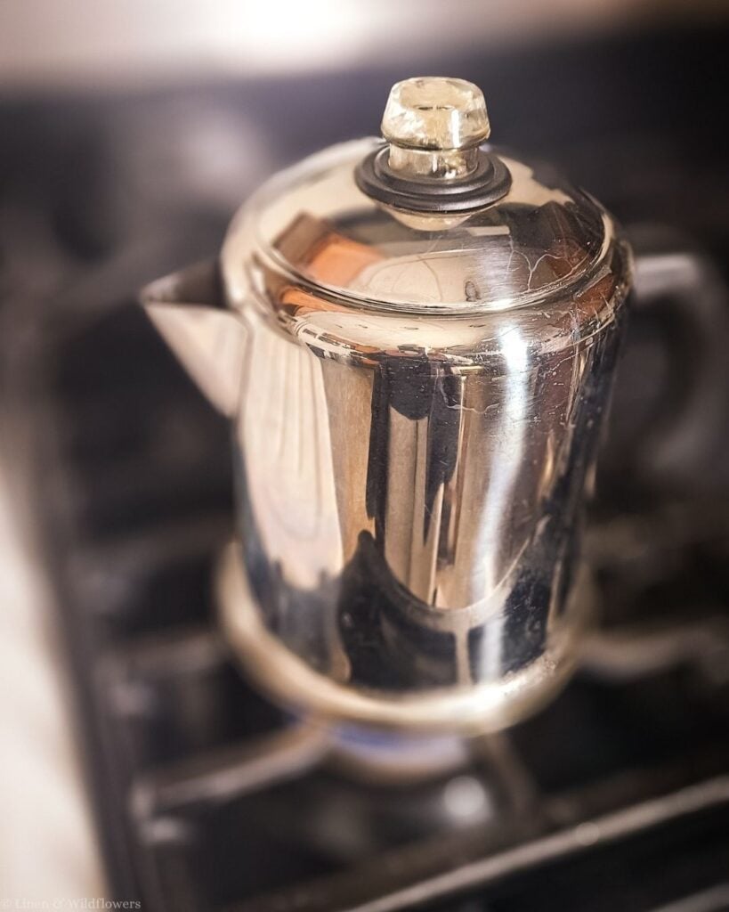 Close-up of a polished stainless steel stovetop percolator with a glass knob lid on the stove.