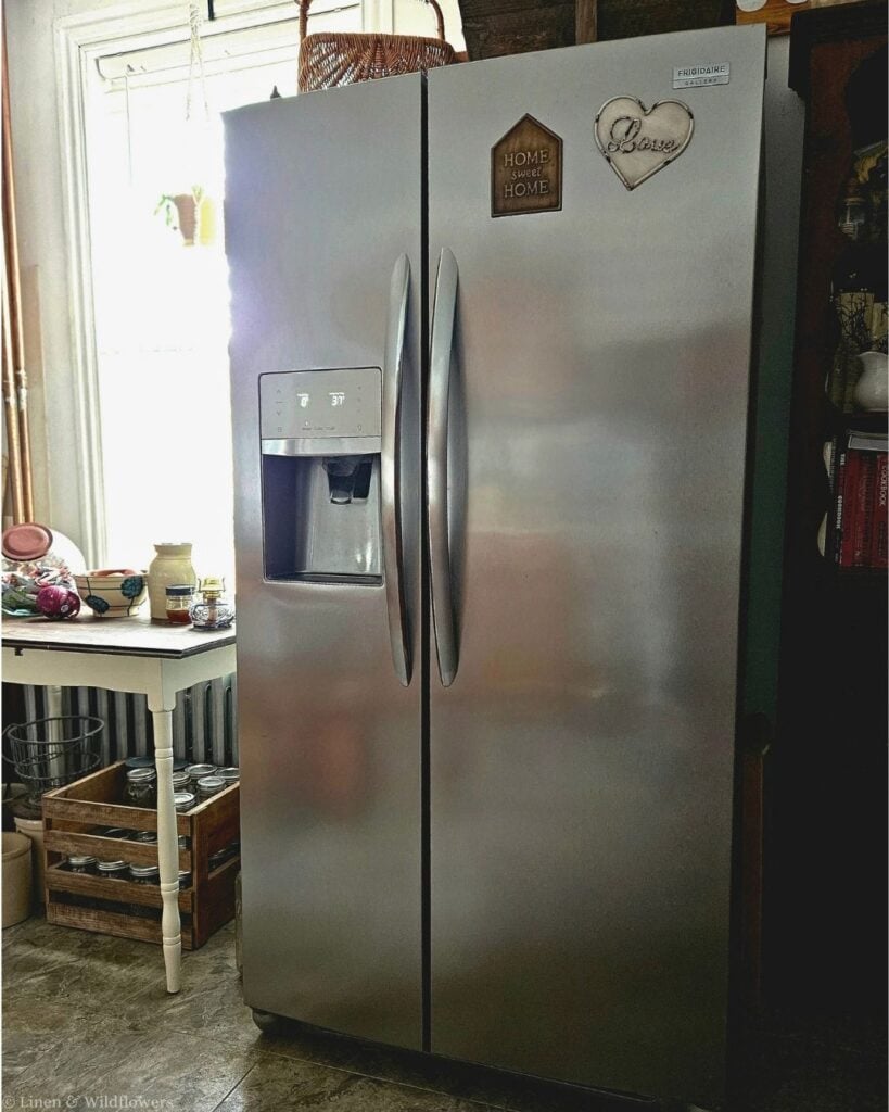 Stainless steel side-by-side refrigerator in a farmhouse kitchen with a crate of canning jars and a vintage table nearby.