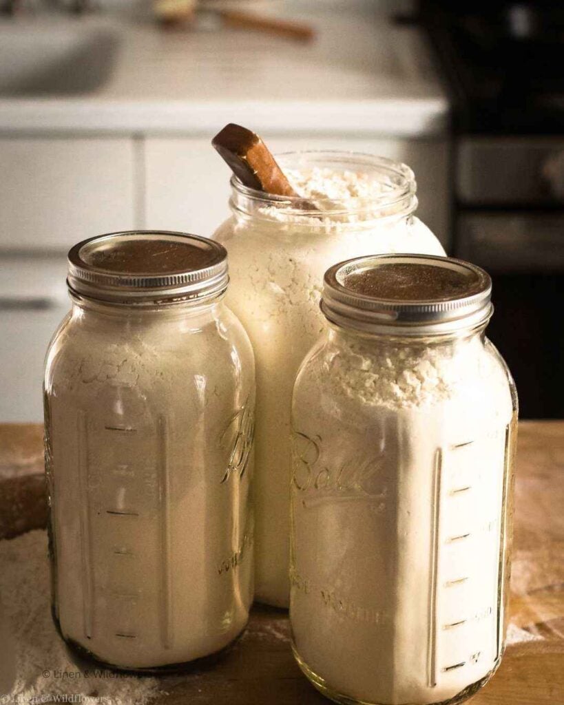 Three Ball wide mouth half gallon mason jars filled with flour and a wooden scoop on a butcher block counter.