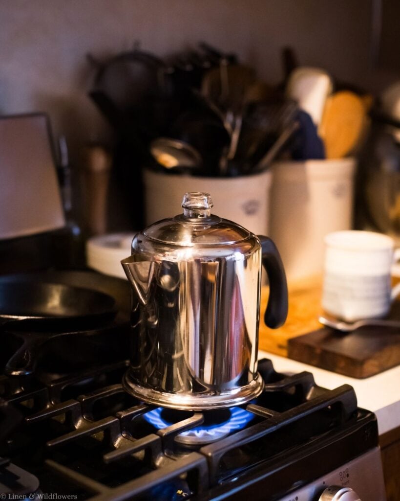 Stainless steel stovetop percolator brewing coffee on a gas burner with a blue flame.
