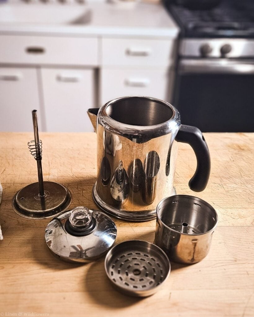 Stovetop percolator taken apart showing the pot, stem, basket, lid and filter on a butcher block counter.