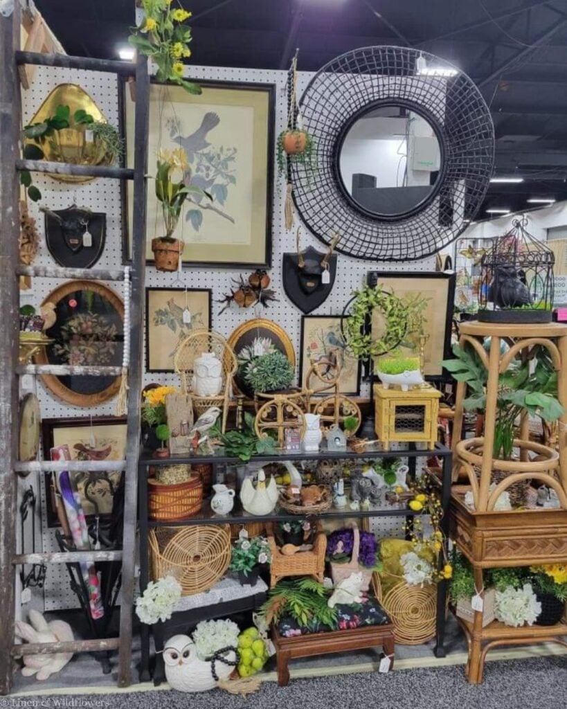 Antique market booth with framed bird prints, rattan pieces, a round black mirror, and vintage owl figurines.