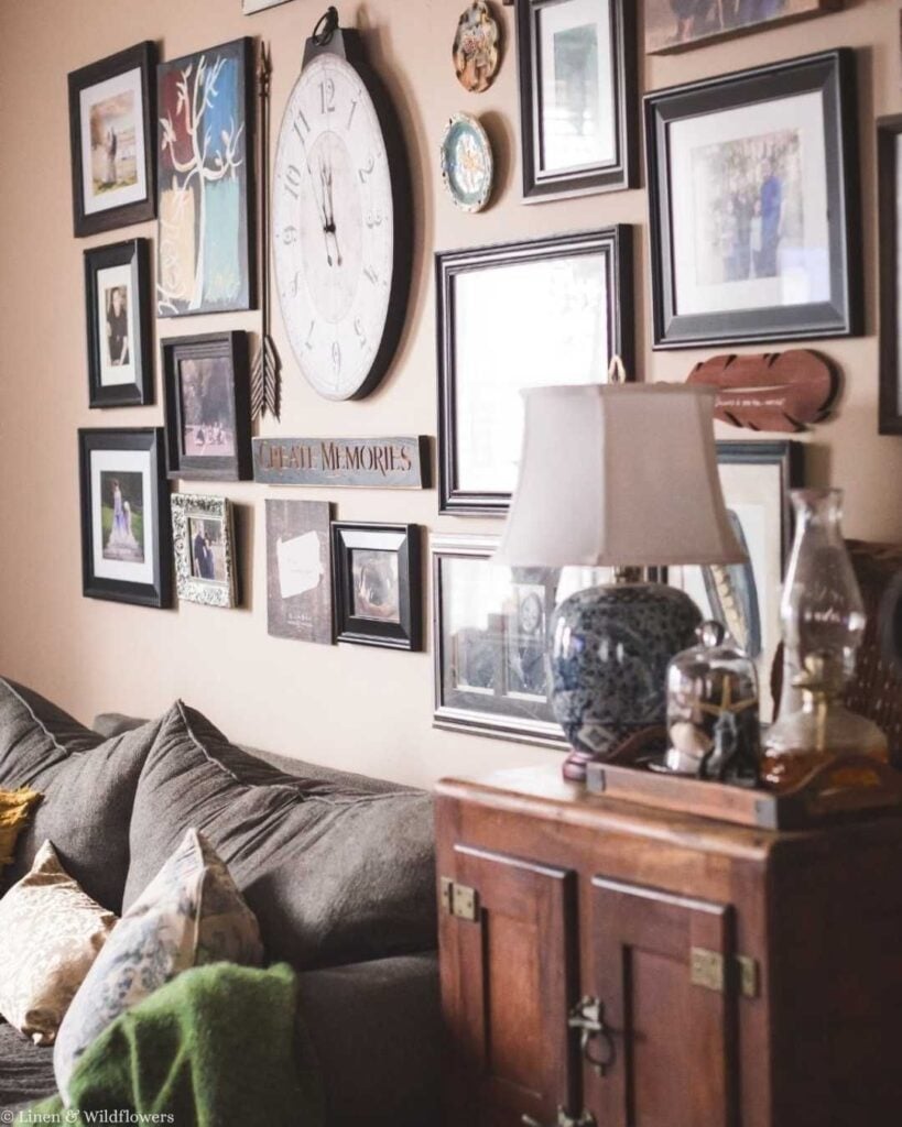 Eclectic gallery wall with framed photos and a large clock above an antique wood cabinet topped with a lamp.