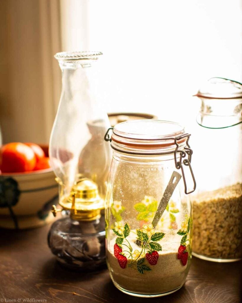 Vintage strawberry glass clamp jar of flour next to a jar of oats and an oil lamp in a sunny kitchen window.
