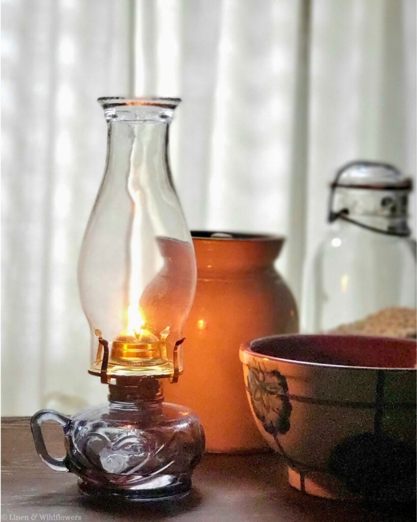 Lit vintage oil lamp beside a stoneware crock, a vintage mixing bowl and a glass canning jar in cozy kitchen window light.