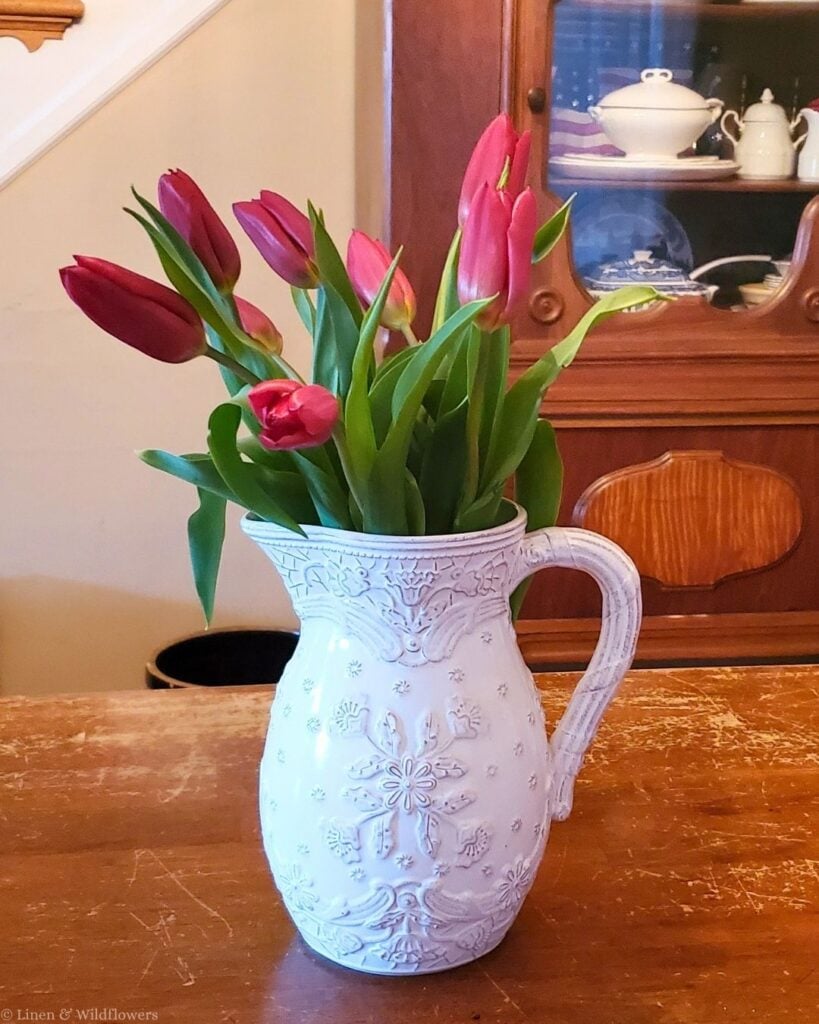 White embossed pitcher filled with red tulips on a wood table in front of an antique hutch with ironstone.