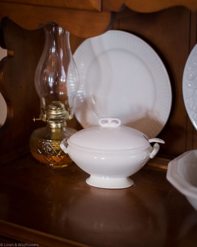 White ironstone soup tureen with ladle beside a glass oil lamp and ironstone plates on a wooden hutch shelf.